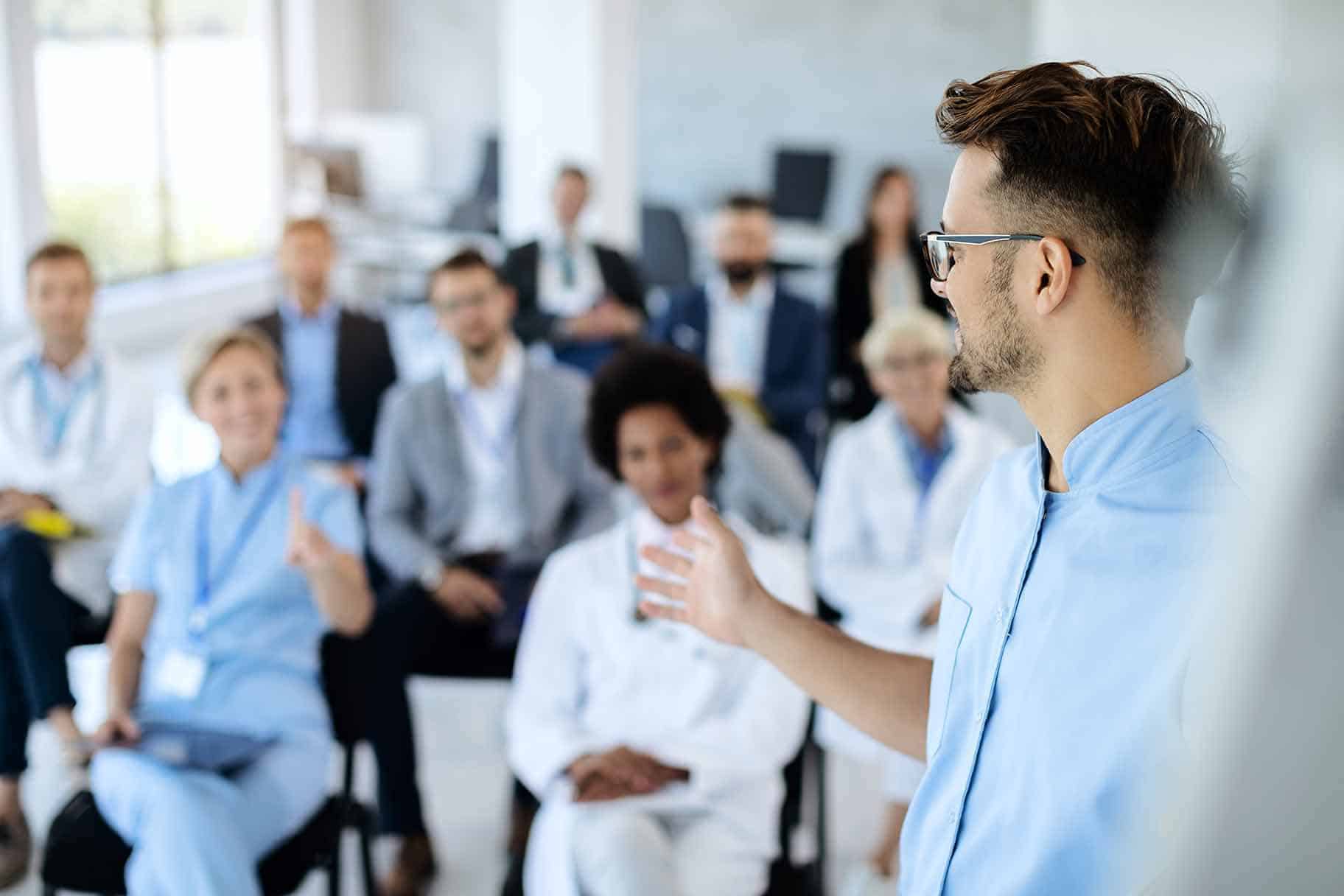 Organizational Leadership in Healthcare Master’s Degree Online a healthcare administrator standing and leading a presentation to a group of seated peers